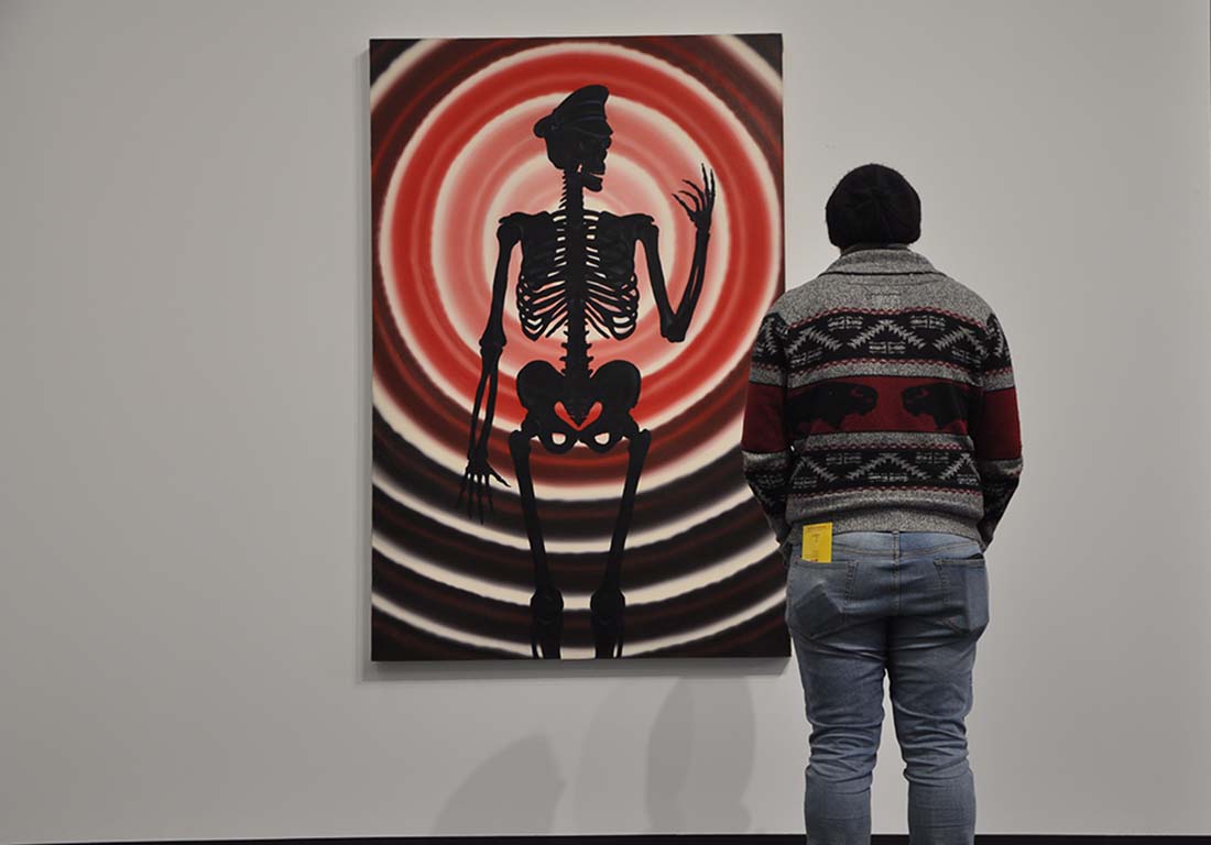 A man stands before a painting of a skeleton against a red target backdrop in the Art AIDs America exhibit.